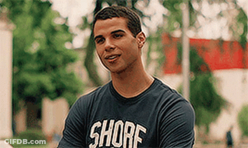 A Young Man Wearing A Shore T-Shirt Is Smiling While Standing In A Park . GIF