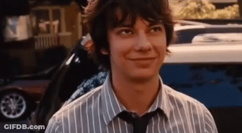 A Young Man Wearing A Striped Shirt And Tie Is Smiling While Standing In Front Of A Car . GIF