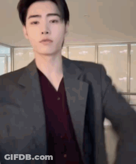 A Young Man Wearing A Suit And A Red Shirt Is Standing In Front Of A Window . GIF