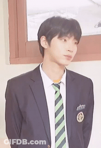 A Young Man Wearing A Suit And Tie Stands In Front Of A Window . GIF