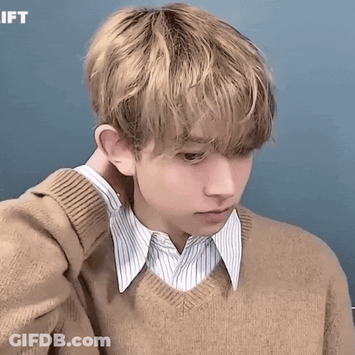 A Young Man Wearing A Tan Sweater And A Striped Shirt Has His Hand On His Neck GIF