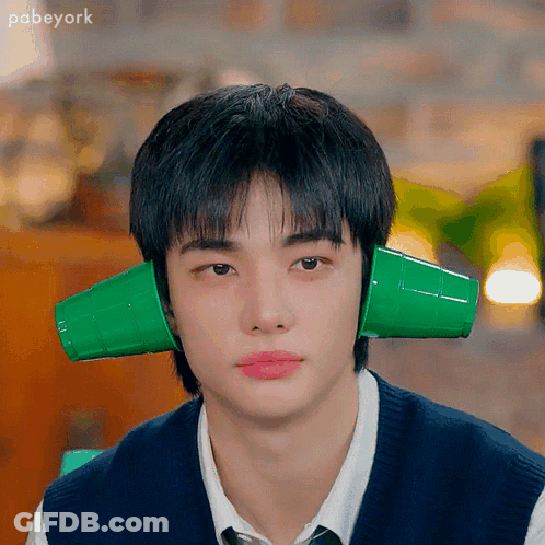 A Young Man Wearing A Vest And Tie Has Two Green Cups On His Ears . GIF