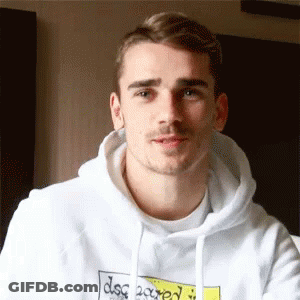 A Young Man Wearing A White Hoodie With A Yellow Sticker On It Is Looking At The Camera . GIF