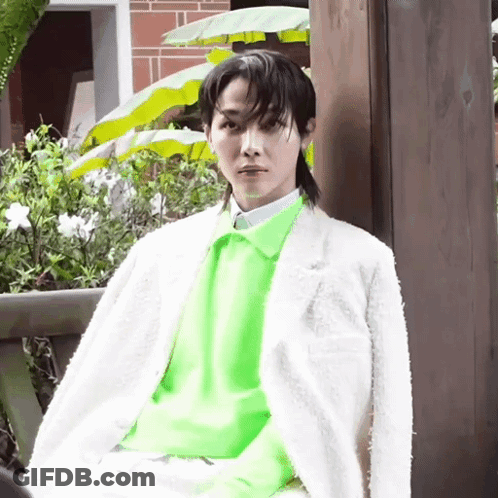 A Young Man Wearing A White Jacket And A Neon Green Sweater Is Sitting On A Bench . GIF