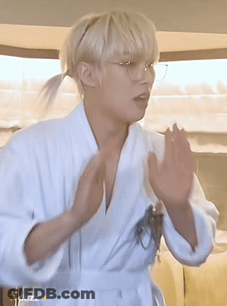 A Young Man Wearing A White Robe And Glasses Is Standing In A Room . GIF