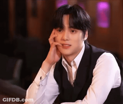 A Young Man Wearing A White Shirt And A Black Vest Is Sitting Down With His Hand On His Face . GIF