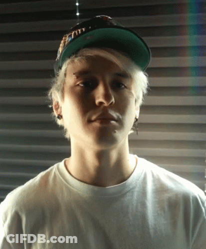 A Young Man Wearing A White Shirt And A Green Hat Looks At The Camera With A Serious Look On His Face GIF