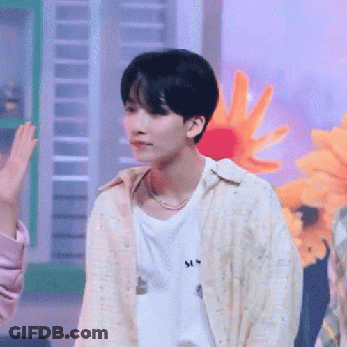 A Young Man Wearing A White Shirt And A Pearl Necklace Is Standing In Front Of A Bunch Of Flowers . GIF