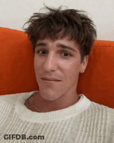 A Young Man Wearing A White Sweater Is Laying On An Orange Pillow GIF