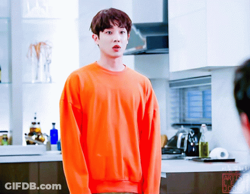 A Young Man Wearing An Orange Sweater Stands In A Kitchen GIF