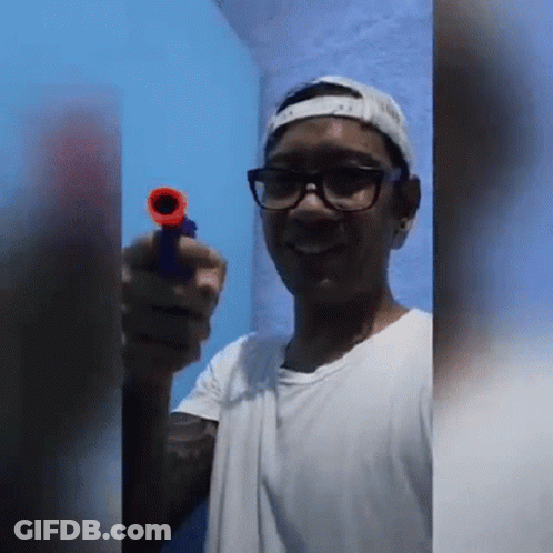 A Young Man Wearing Glasses And A Baseball Cap Is Holding A Toy Gun . GIF