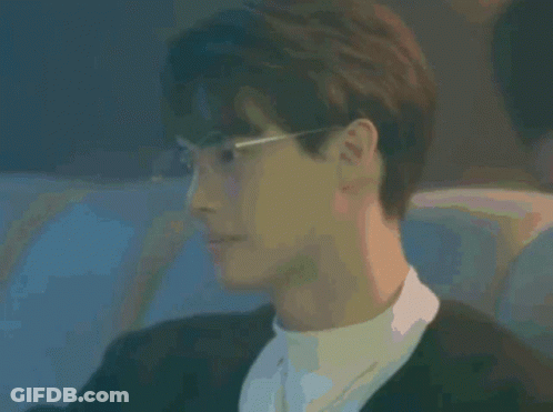 A Young Man Wearing Glasses And A Black Jacket Is Sitting In A Dark Room . GIF
