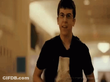 A Young Man Wearing Glasses And A Black Shirt Is Holding A Piece Of Paper In A Hallway . GIF