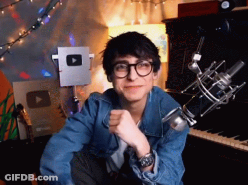 A Young Man Wearing Glasses And A Denim Jacket Is Sitting In Front Of A Microphone And A Piano . GIF