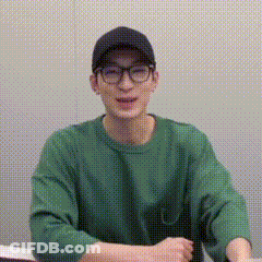 A Young Man Wearing Glasses And A Green Sweater Is Sitting At A Table . GIF