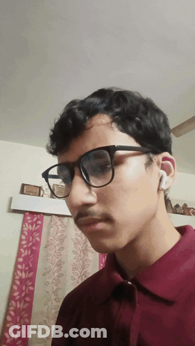 A Young Man Wearing Glasses And A Maroon Shirt Is Looking At The Camera GIF