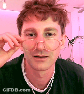 A Young Man Wearing Glasses And A Necklace Is Looking At The Camera . GIF