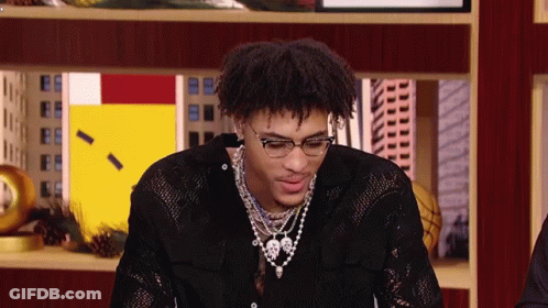 A Young Man Wearing Glasses And A Necklace Is Sitting In Front Of A Display Case . GIF