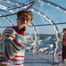 A Young Man Wearing Glasses And A Striped Sweater Sings Into A Microphone GIF