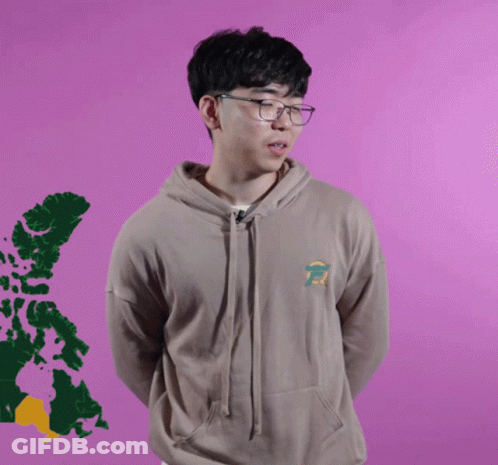 A Young Man Wearing Glasses And A Tan Hoodie Stands In Front Of A Purple Background GIF