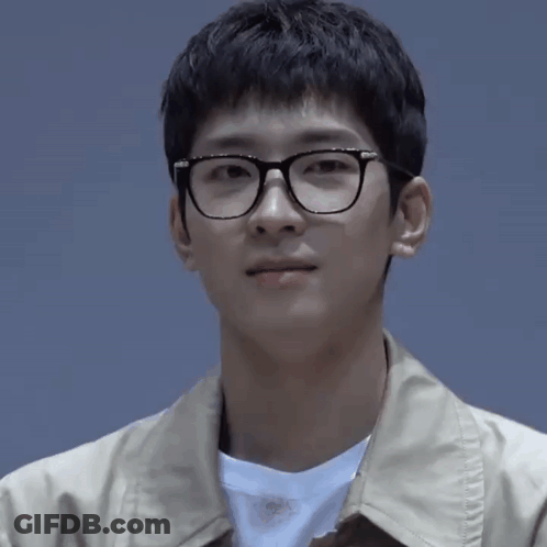 A Young Man Wearing Glasses And A Tan Jacket Is Looking At The Camera GIF