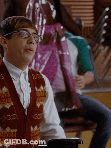 A Young Man Wearing Glasses And A Vest That Says ' A ' On It Sits In A Chair GIF