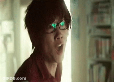 A Young Man Wearing Glasses Is Making A Surprised Face In A Library . GIF