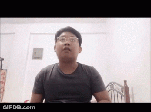 A Young Man Wearing Glasses Is Sitting In Front Of A Bed . GIF