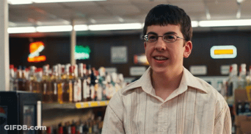 A Young Man Wearing Glasses Is Smiling In Front Of A Shelf Of Liquor GIF