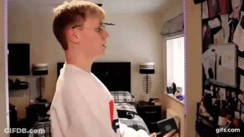 A Young Man Wearing Glasses Is Standing In A Bedroom Next To A Bed And A Window . GIF