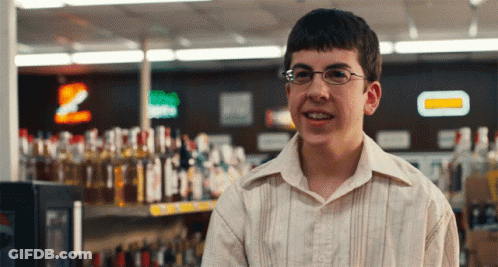 A Young Man Wearing Glasses Stands In Front Of A Shelf Of Liquor Bottles GIF