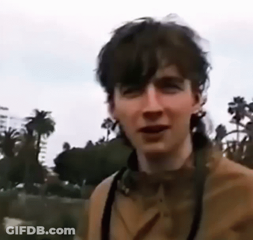 A Young Man Wearing Headphones And A Camera Is Standing In Front Of Palm Trees . GIF