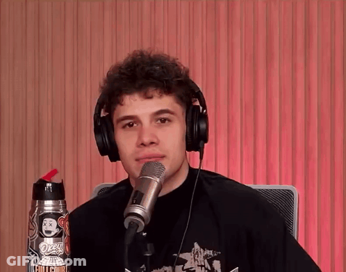 A Young Man Wearing Headphones Stands In Front Of A Microphone With A Sticker On His Shirt That Says Ozzy GIF