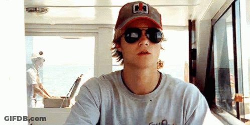 A Young Man Wearing Sunglasses And A Baseball Cap Is Sitting On A Boat . GIF