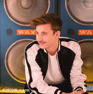 A Young Man With A Mustache Is Sitting In Front Of A Wall Of Speakers With The Word Wax On It . GIF