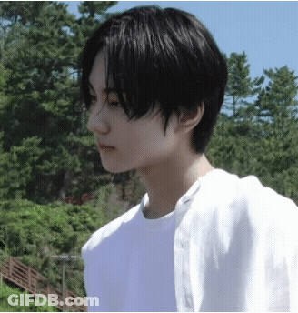 A Young Man With Black Hair And A White Shirt Is Standing In Front Of Trees GIF