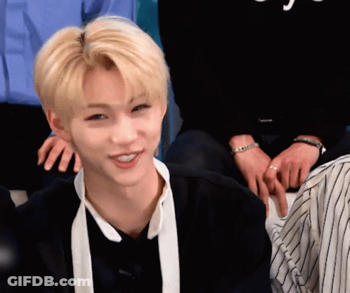 A Young Man With Blonde Hair And A White Scarf Around His Neck Is Smiling While Sitting In A Room With Other People . GIF