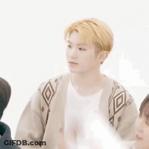 A Young Man With Blonde Hair Is Sitting In Front Of A White Wall With His Arm In The Air . GIF