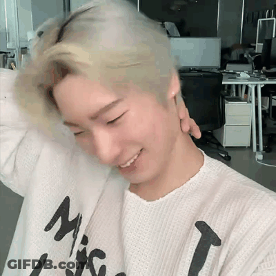 A Young Man With Blonde Hair Is Wearing A White Sweater With A Black Letter T On It . GIF