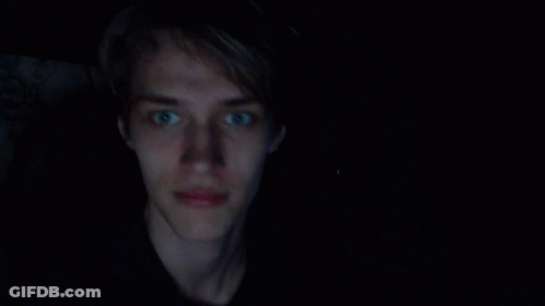 A Young Man With Blue Eyes Is Smiling In A Dark Room GIF