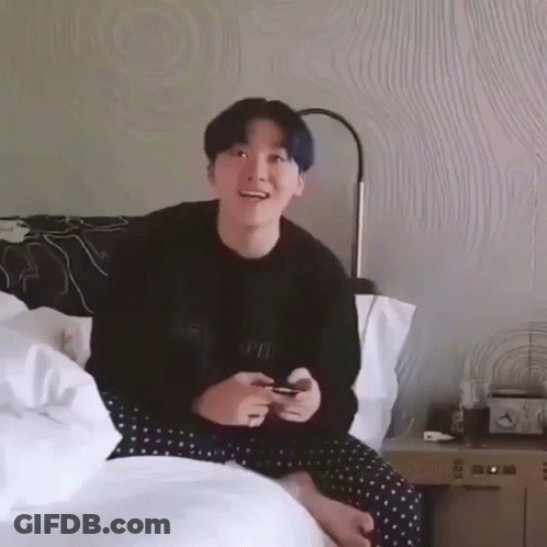 A Young Man With Blue Hair Is Sitting On A Bed Playing A Video Game GIF