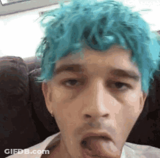 A Young Man With Blue Hair Is Sticking His Tongue Out While Sitting On A Couch . GIF