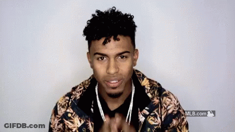 A Young Man With Curly Hair And A Beard Is Wearing A Floral Jacket And Necklace . GIF