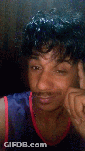 A Young Man With Curly Hair And A Mustache Is Wearing A Blue And Red Tank Top GIF
