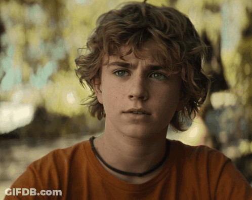 A Young Man With Curly Hair And A Necklace Around His Neck Looks At The Camera GIF