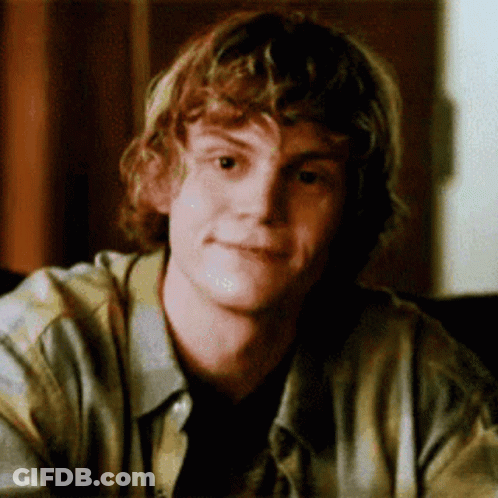 A Young Man With Curly Hair Is Sitting At A Table Looking At The Camera . GIF