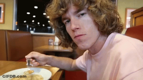 A Young Man With Curly Hair Is Sitting At A Table With A Plate Of Food And A Fork In His Hand . GIF