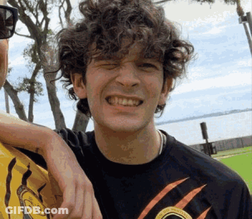 A Young Man With Curly Hair Is Smiling For The Camera While Standing Next To Another Man . GIF