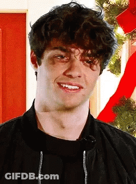 A Young Man With Curly Hair Is Smiling In Front Of A Red Christmas Tree GIF