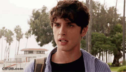 A Young Man With Curly Hair Is Standing In Front Of A Lifeguard Tower And Palm Trees . GIF